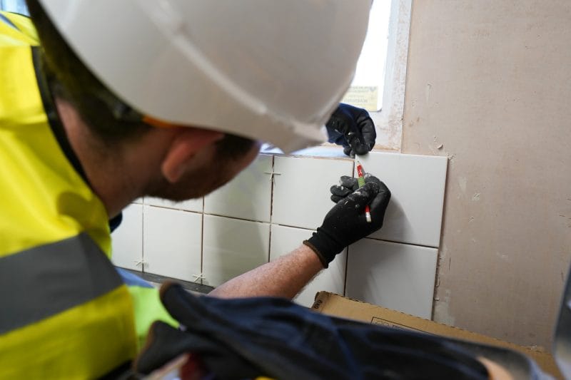 Axis CLC Group worker in a hard hat and high-vis vest carefully measuring and fitting wall tiles.