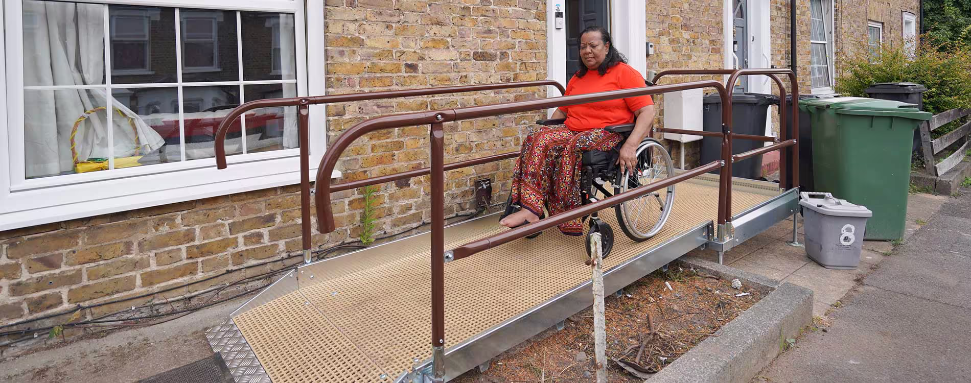A female resident in a wheelchair uses a newly installed, custom-built metal ramp to safely access the front door of her brick home.