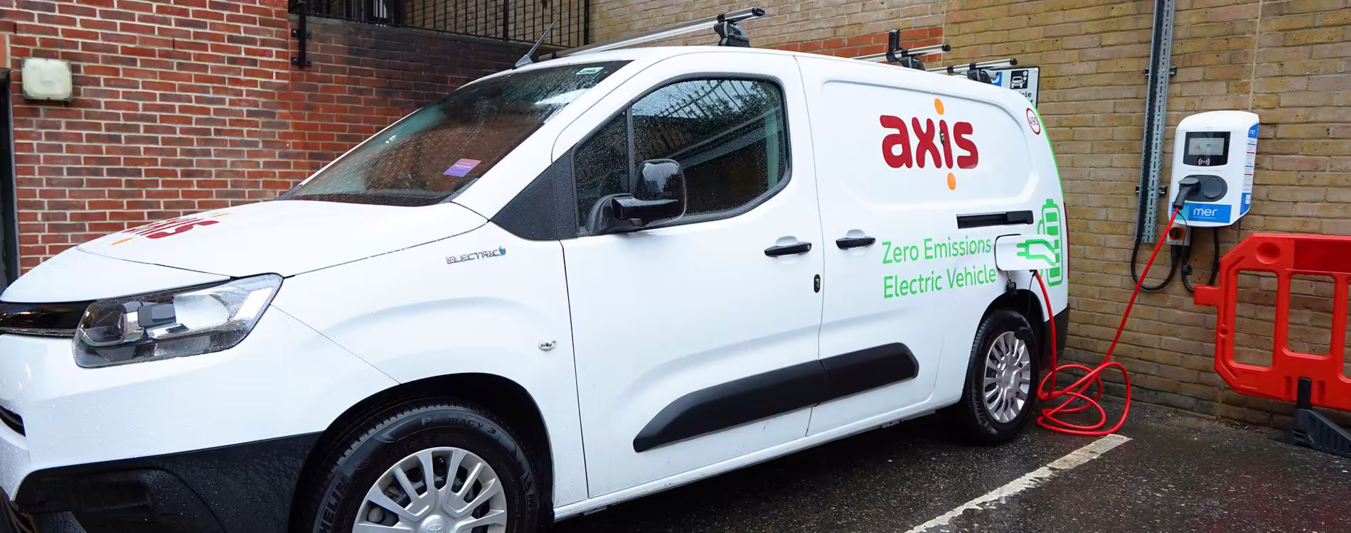 An Axis-branded 'Zero Emissions Electric Vehicle' van is parked and plugged into a wall-mounted Mer charging station, showcasing a commercial EV fleet installation.