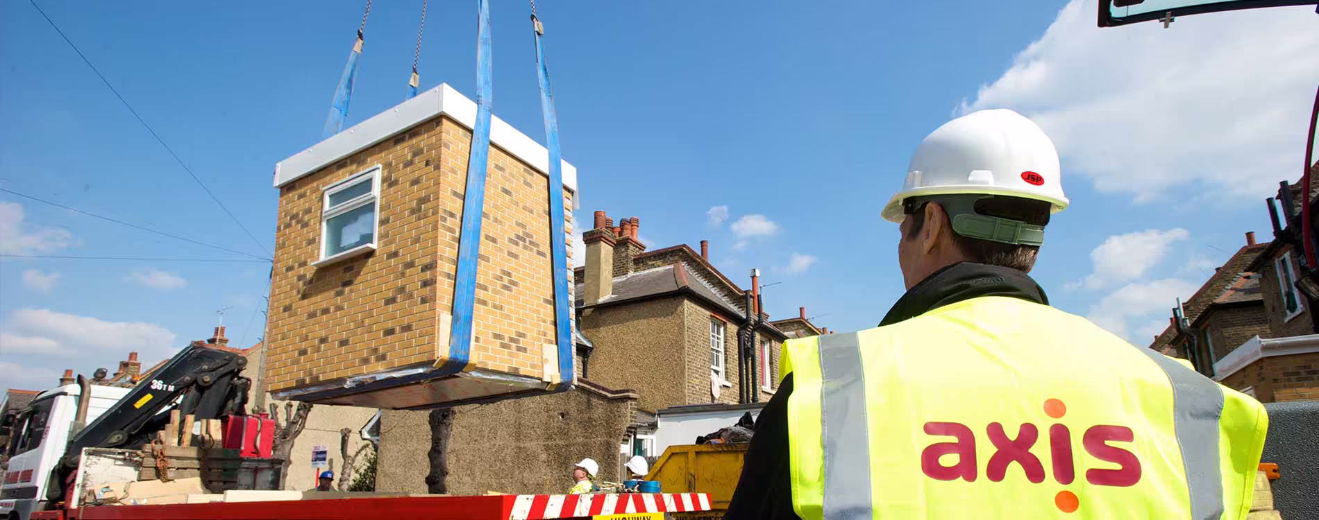 An Axis site manager in a hard hat and hi-vis vest oversees a crane carefully lifting a prefabricated brick bathroom extension pod into place behind a terraced house.