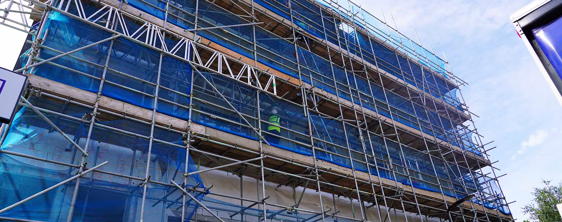 The facade of a large building undergoing cladding remediation works, completely covered in scaffolding and blue safety netting.