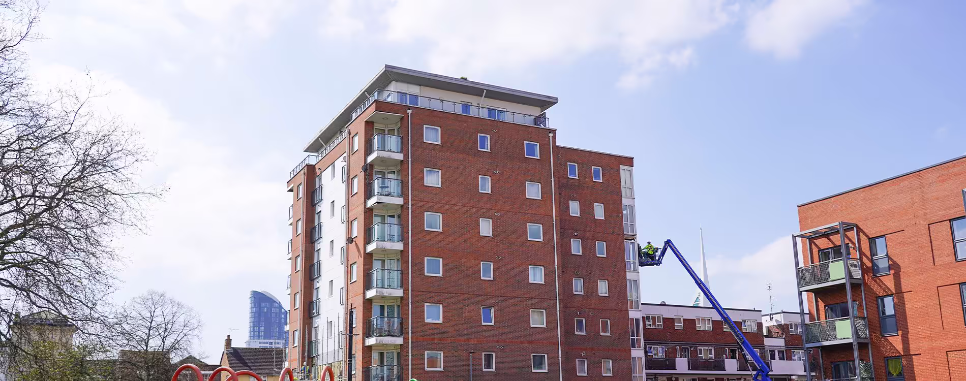 A wide view of a modern red brick apartment block, with an operative in a cherry picker working on the facade during a cladding systems replacement project.