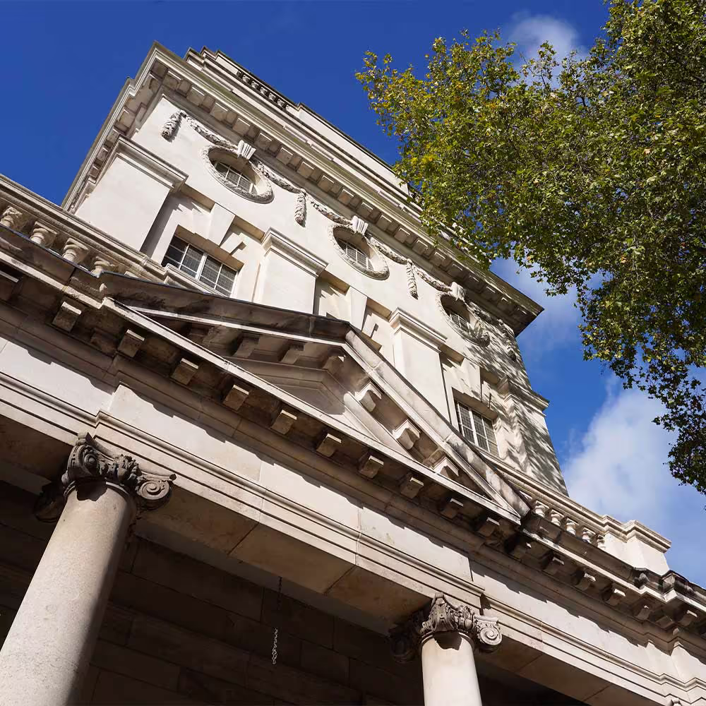 A low-angle view looking up at the grand, classical stone facade of a heritage building, highlighting the ornate columns and stonework against a blue sky.
