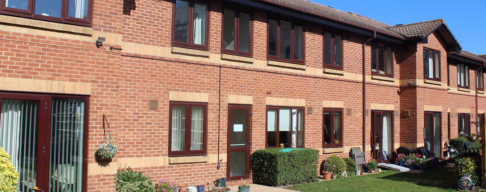The rear exterior of a modern red brick residential building, showing the ground floor patios and gardens after the completion of an exterior home repairs project.