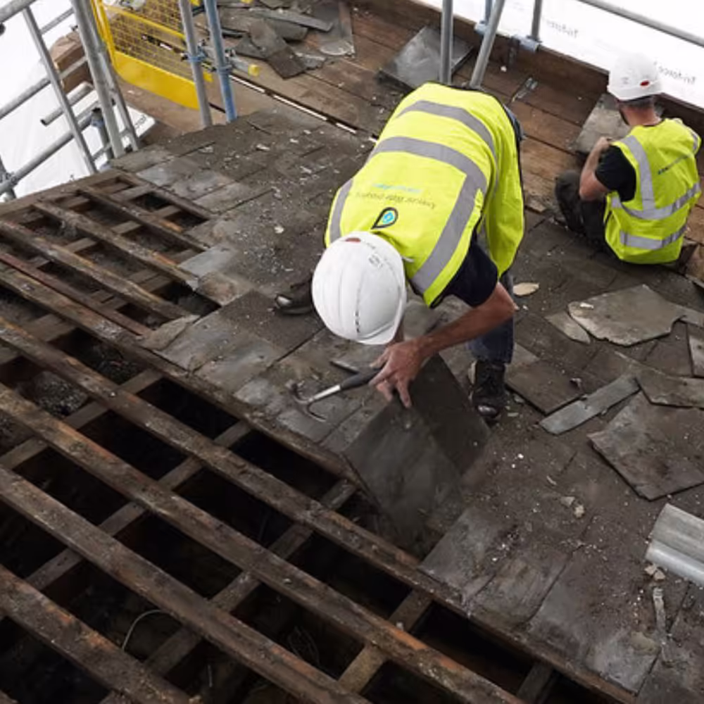 Two heritage roofing specialists in safety gear carefully removing old slates from the roof of the Grade II listed building during its refurbishment.