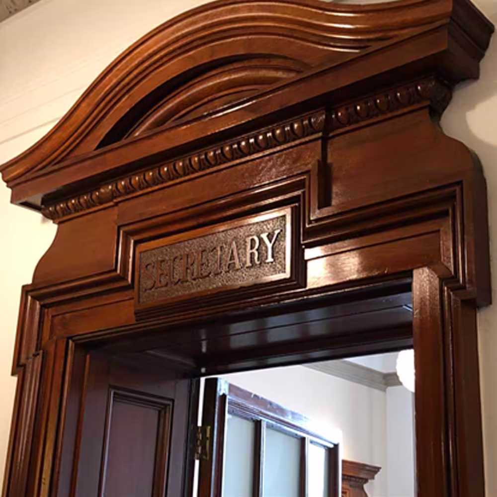 The ornate, restored wooden door frame for the 'Secretary's' office in a Grade II listed building, highlighting the intricate carvings and high-gloss finish.