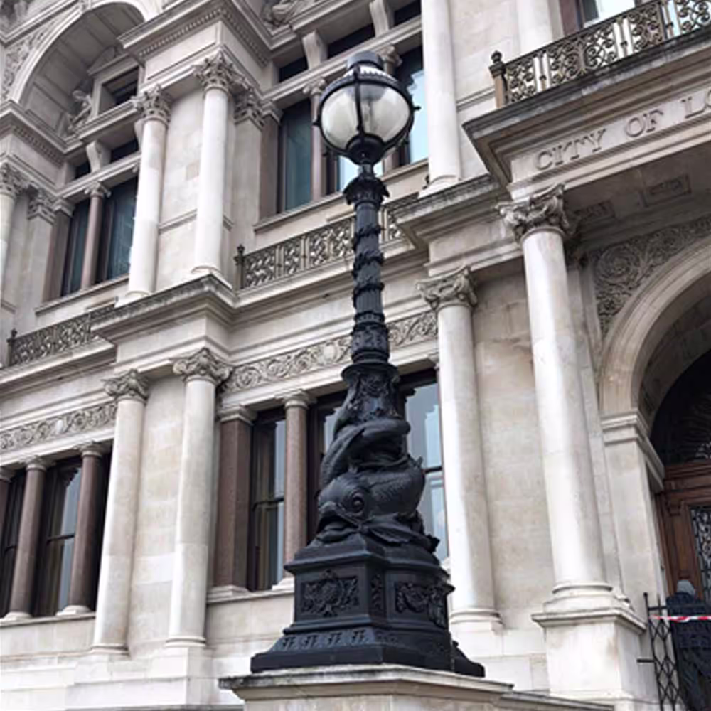 An ornate, historic cast iron lamppost in the City of London, standing before a grand Grade II listed stone building, showcasing heritage street furniture.