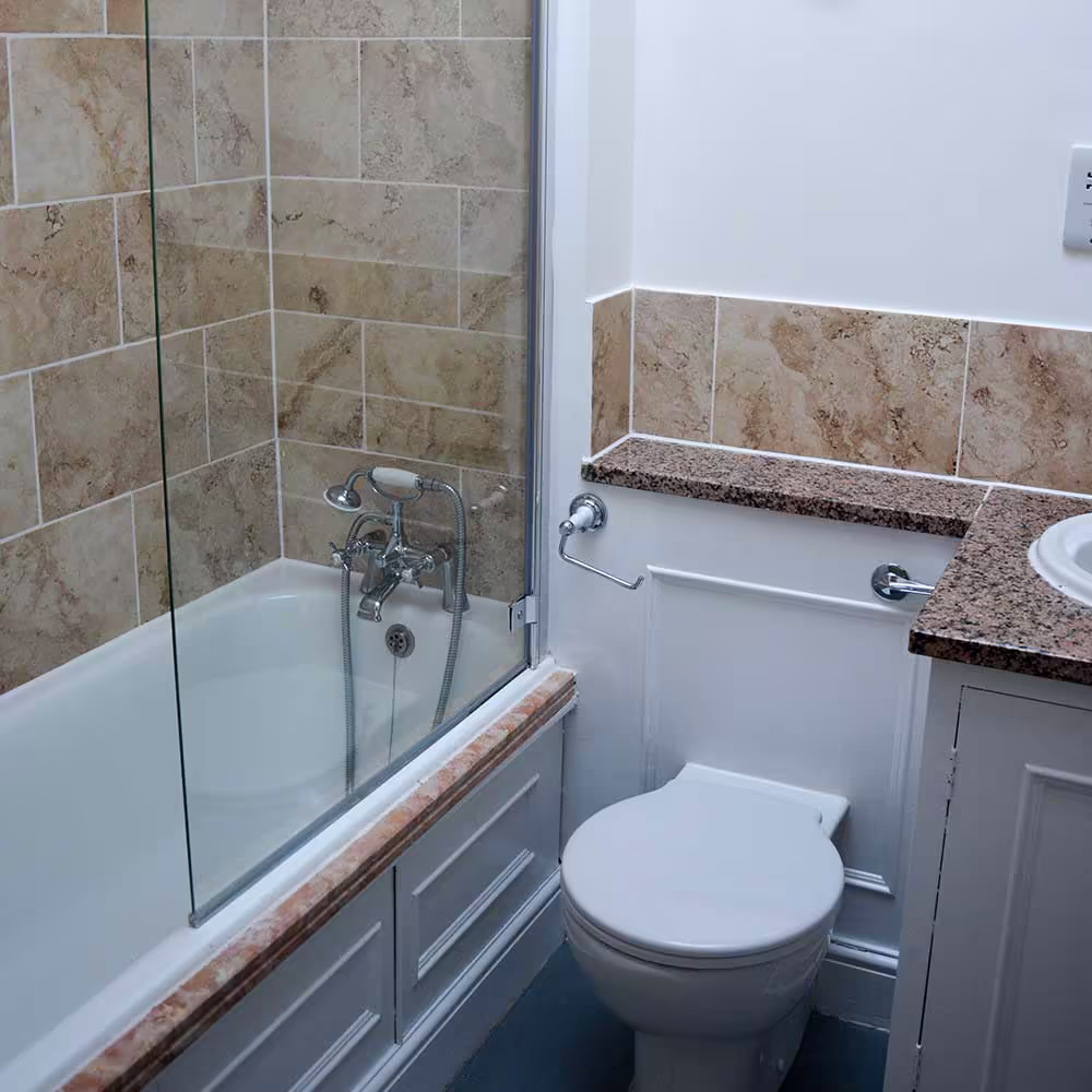 A repaired and modernised bathroom in a Grade Listed property, featuring classic stone-effect tiles, a new toilet, and a bath with a glass shower screen.