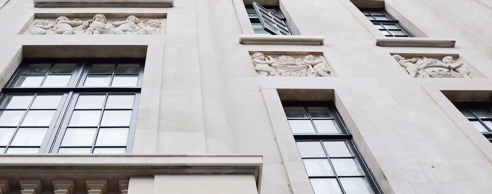 A low-angle view of a conserved heritage building, highlighting the intricate carved stone friezes and panels on the white stone facade above dark-framed windows.