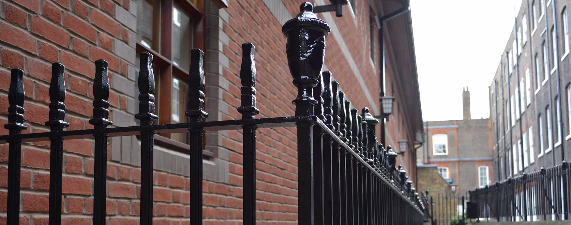 A low-angle view of a beautifully restored heritage cast iron railing, painted in high-gloss black, running alongside a traditional red brick building in London.