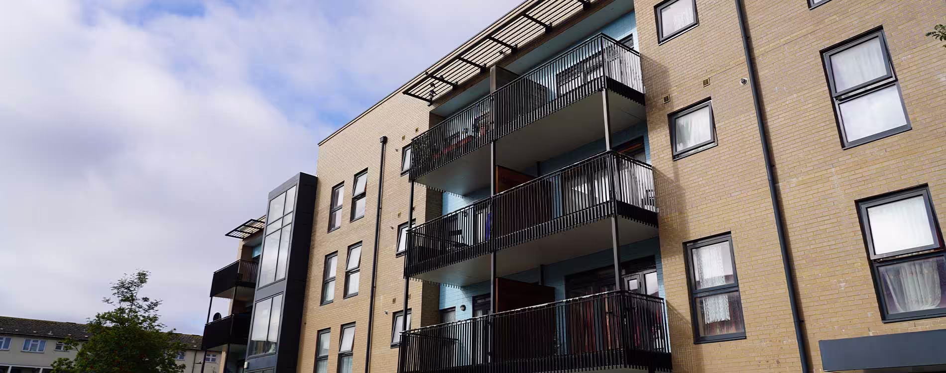 Low-angle shot of a modern PA Housing apartment block with yellow brickwork and stacked black metal balconies, under a partly cloudy sky.