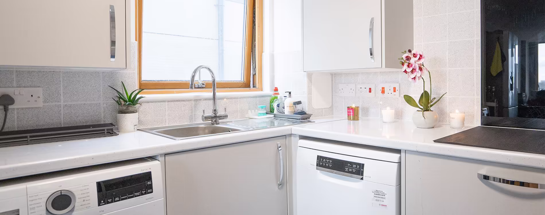 Panoramic view of a bright, modern kitchen after a full refurbishment, showing new light grey units, integrated appliances, and a stainless steel sink.