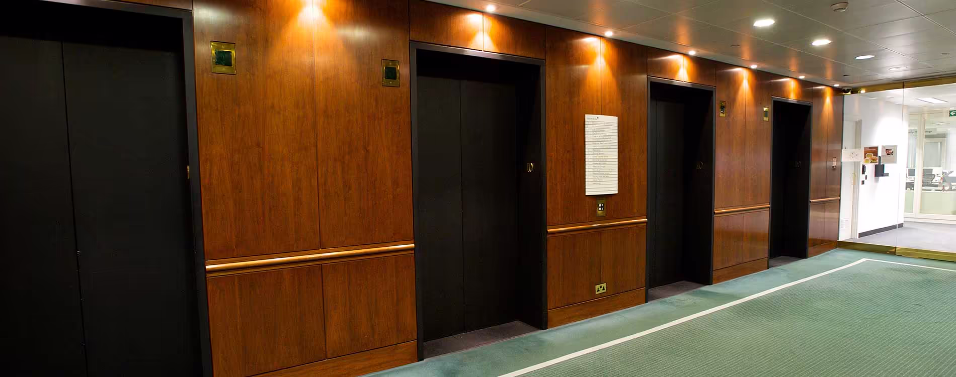 The 'before' view of a lift lobby prior to upgrades, showing traditional dark wood panelling, multiple lifts, and dated green carpet.