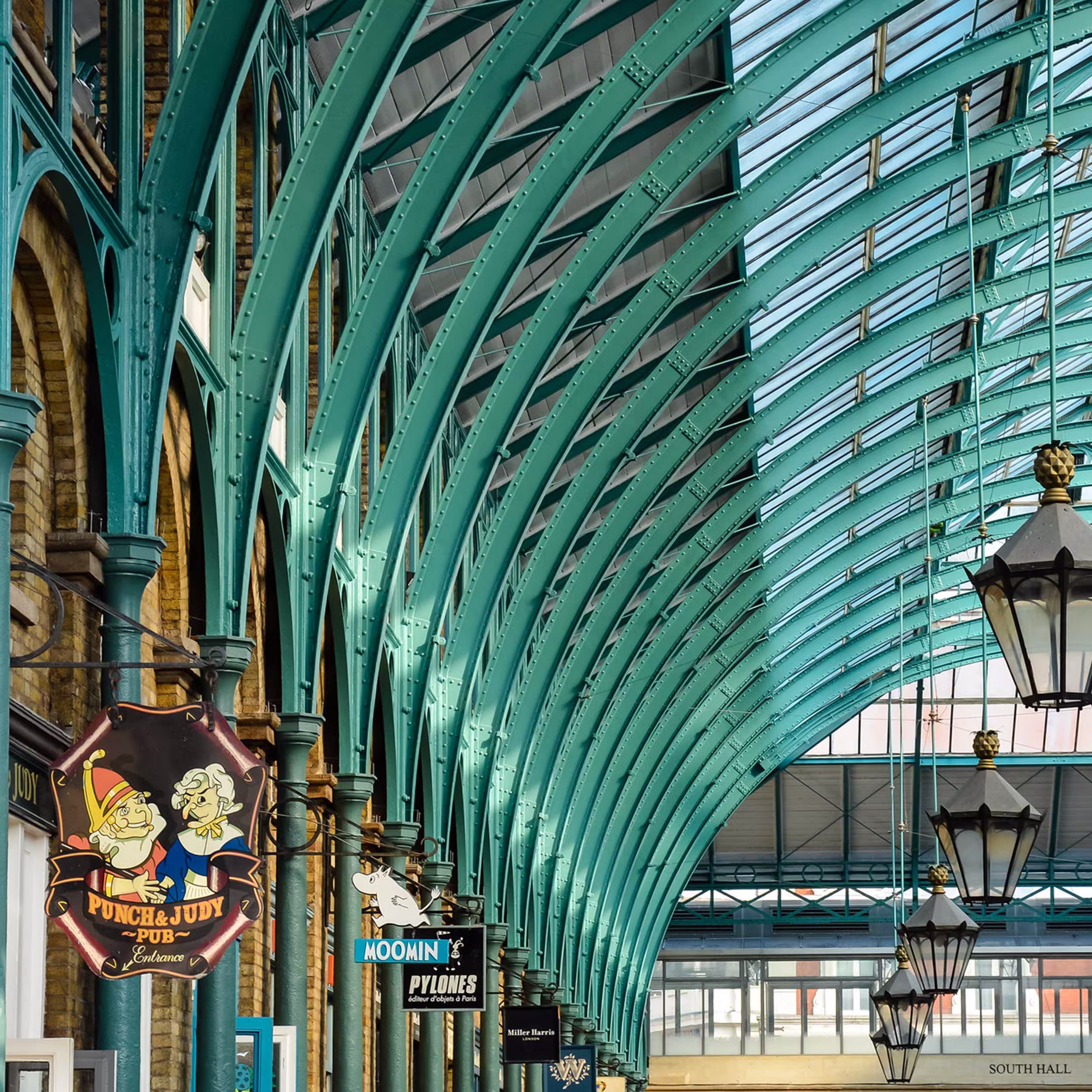 The iconic green arched ironwork of Covent Garden Market's roof, showing the results of the listed building restoration, with traditional lanterns and shop signs.