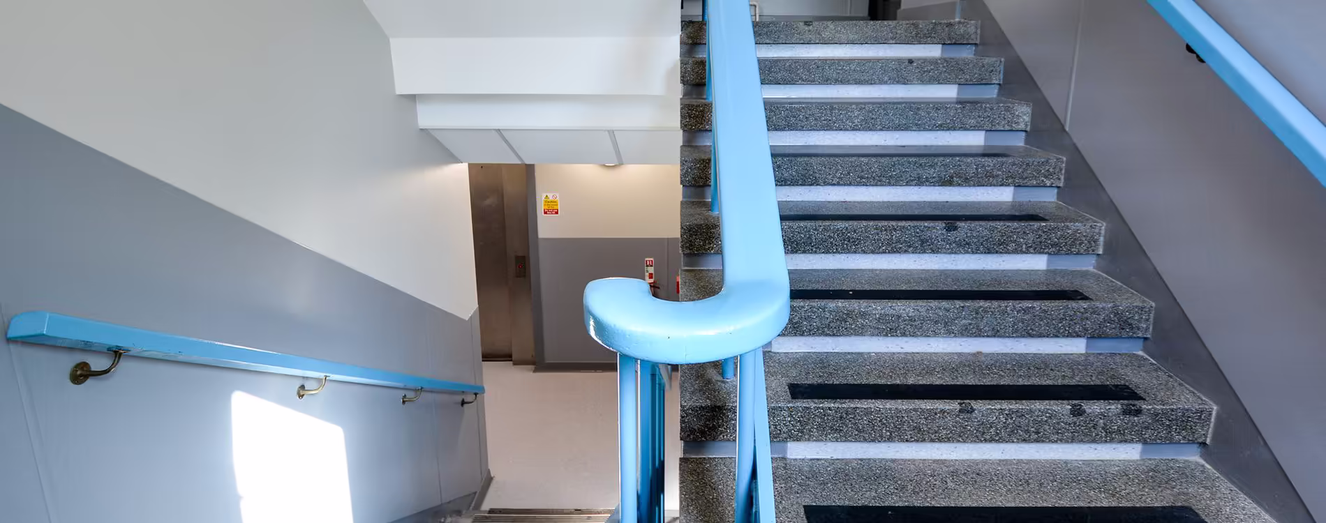 A refurbished communal staircase in a police station, showing the newly painted light blue handrails on the terrazzo-style concrete stairs.