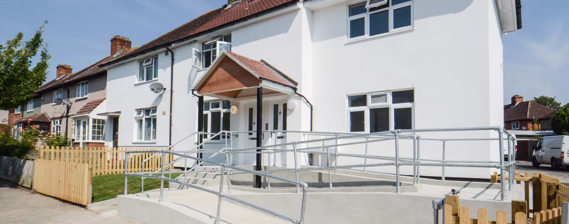A panoramic view of a residential conversion project, showing the newly rendered white building with a large, accessible concrete ramp and handrails leading to the entrance.