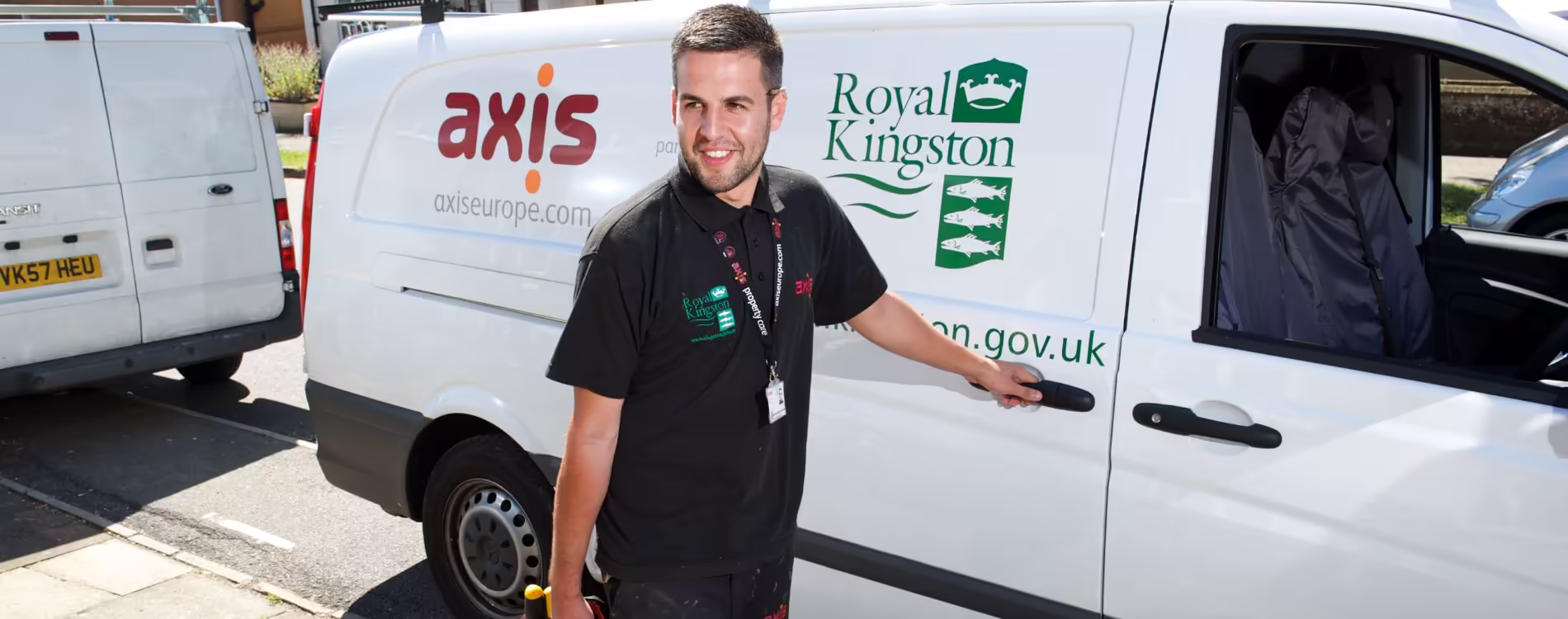 A young, smiling male Axis technician in a black uniform stands next to a white service van, holding the driver's door handle. The van is branded with the Axis logo and the 'Royal Kingston' logo, indicating a responsive repairs service in the Royal Borough of Kingston.