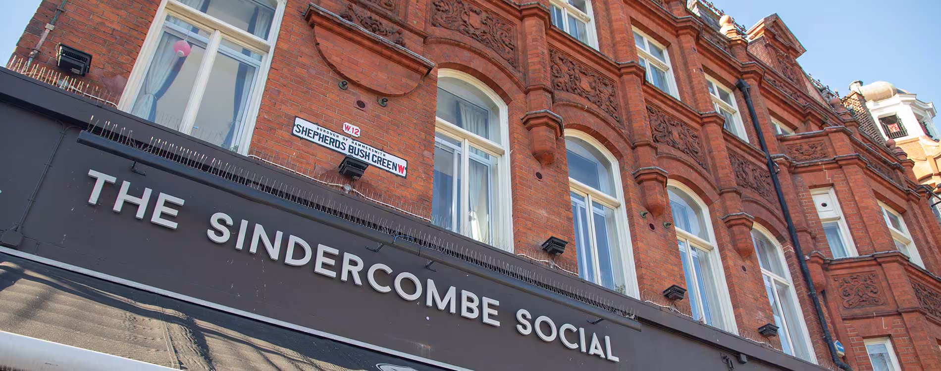 Close-up view of the red brick Victorian building facade above the new signage for 'The Sindercombe Social' pub on Shepherds Bush Green. The sign is black with white lettering.