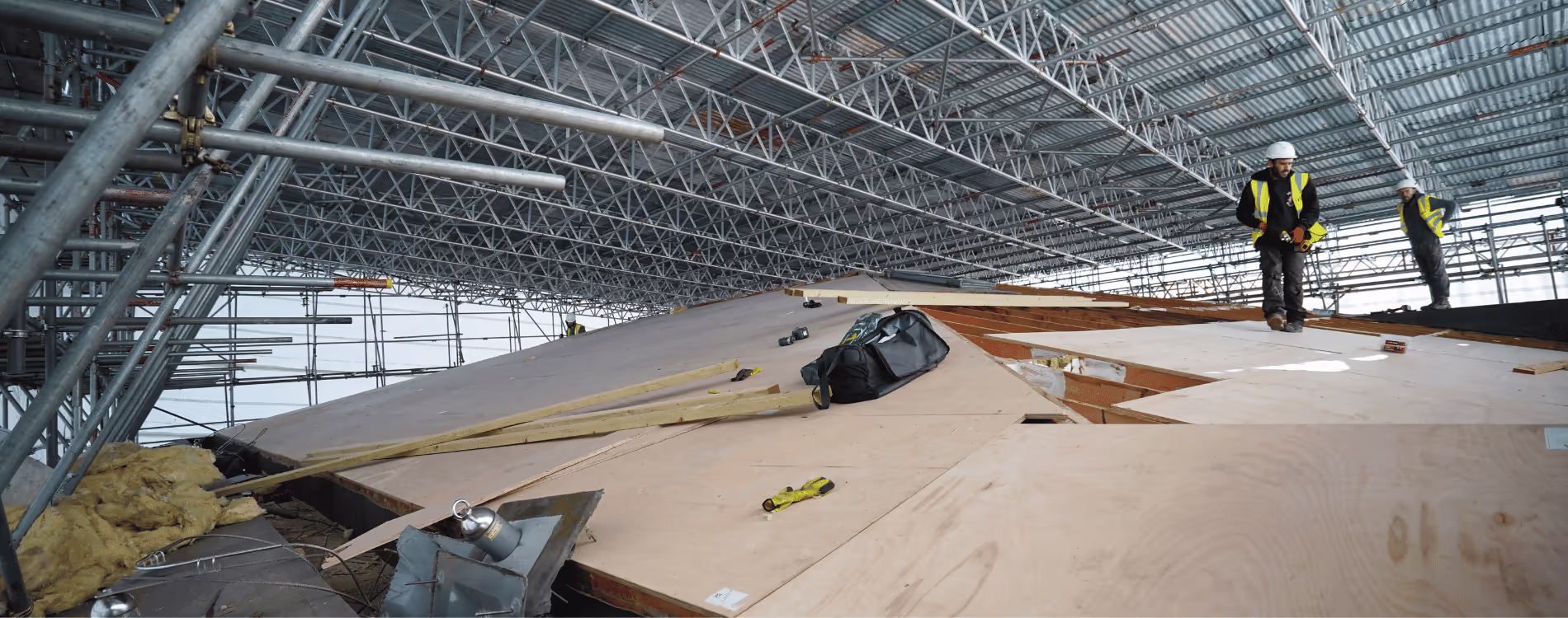 Construction workers in high-visibility vests and hard hats stand on a large, newly boarded timber roof structure under an extensive temporary roof scaffolding system, indicating major roof renewal programme work.