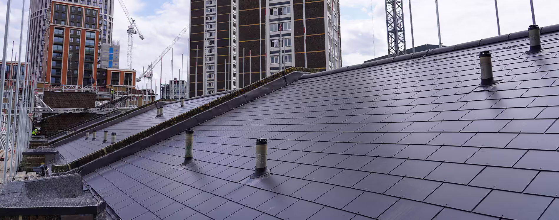 A wide, elevated view of a newly installed, dark grey pitched slate roof with ventilation pipes, set against a backdrop of modern high-rise residential buildings and construction cranes in a city, illustrating a major roof renewal project.