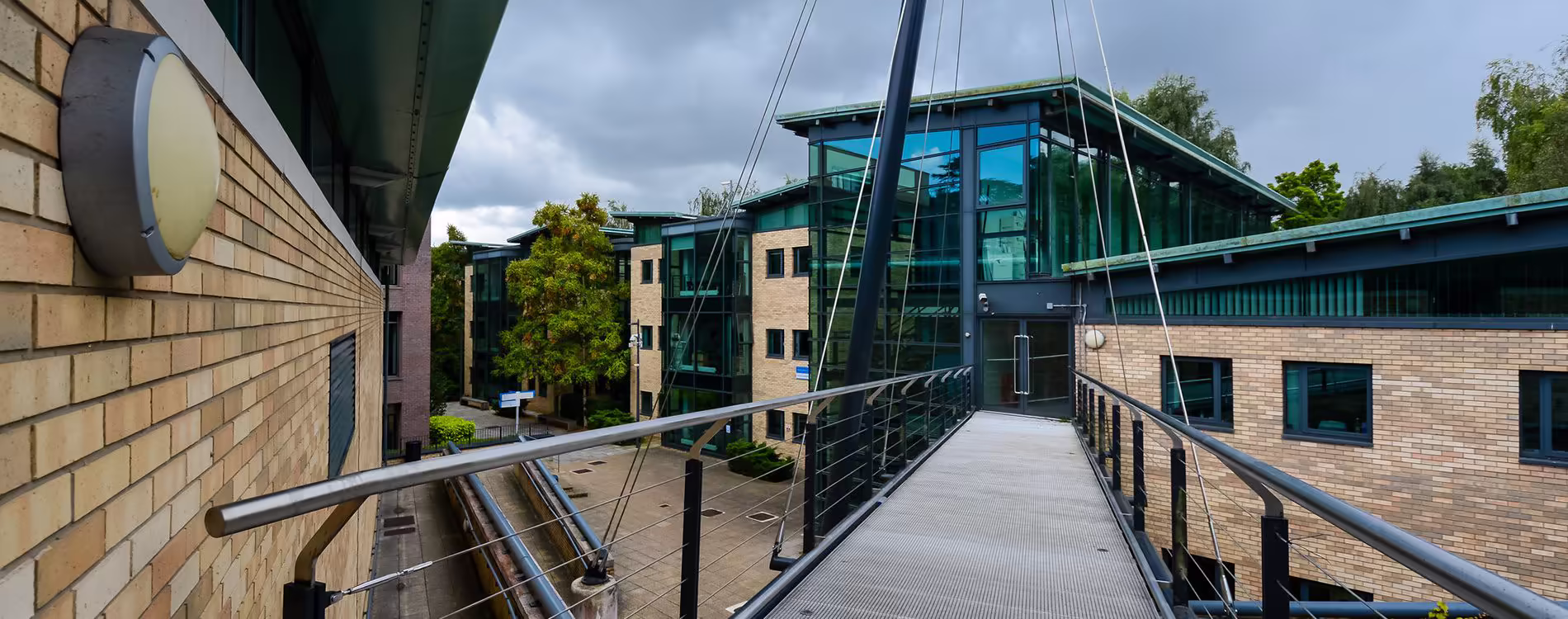 An elevated view of a contemporary university campus, showing a pedestrian bridge or walkway connecting two buildings with modern yellow and dark brick facades and large glass panels, illustrating campus maintenance and access improvements.