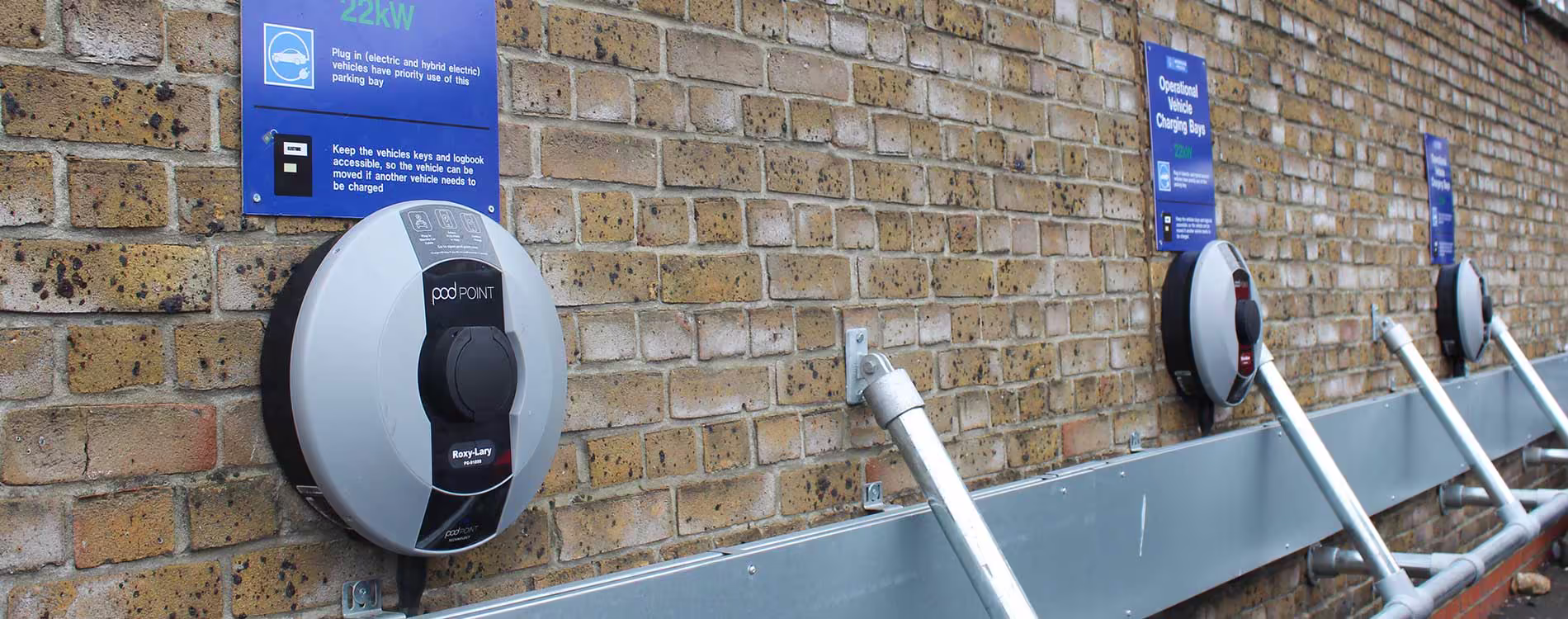 A row of newly installed Pod Point electric vehicle (EV) charging units mounted on a yellow brick wall. Blue signage indicates 'Operational Vehicle Charging Bays' for use by electric and hybrid vehicles.