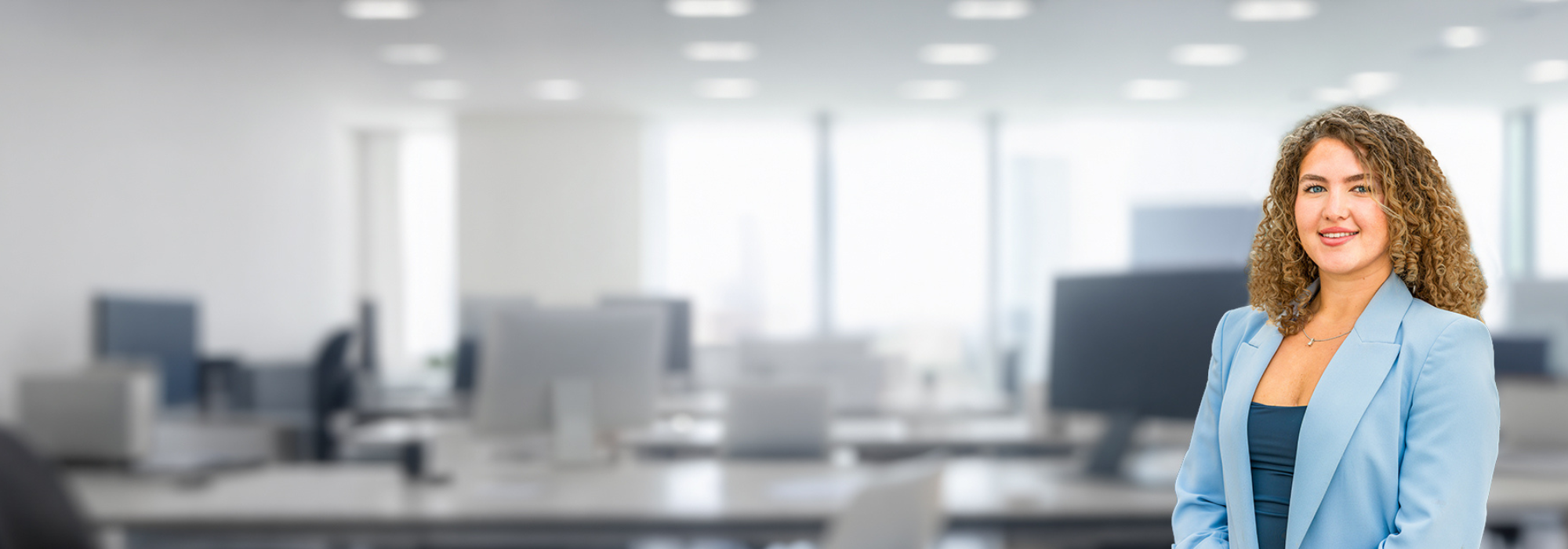 Smiling woman with blue blazer and top. Office background with desk and monitors behind. 