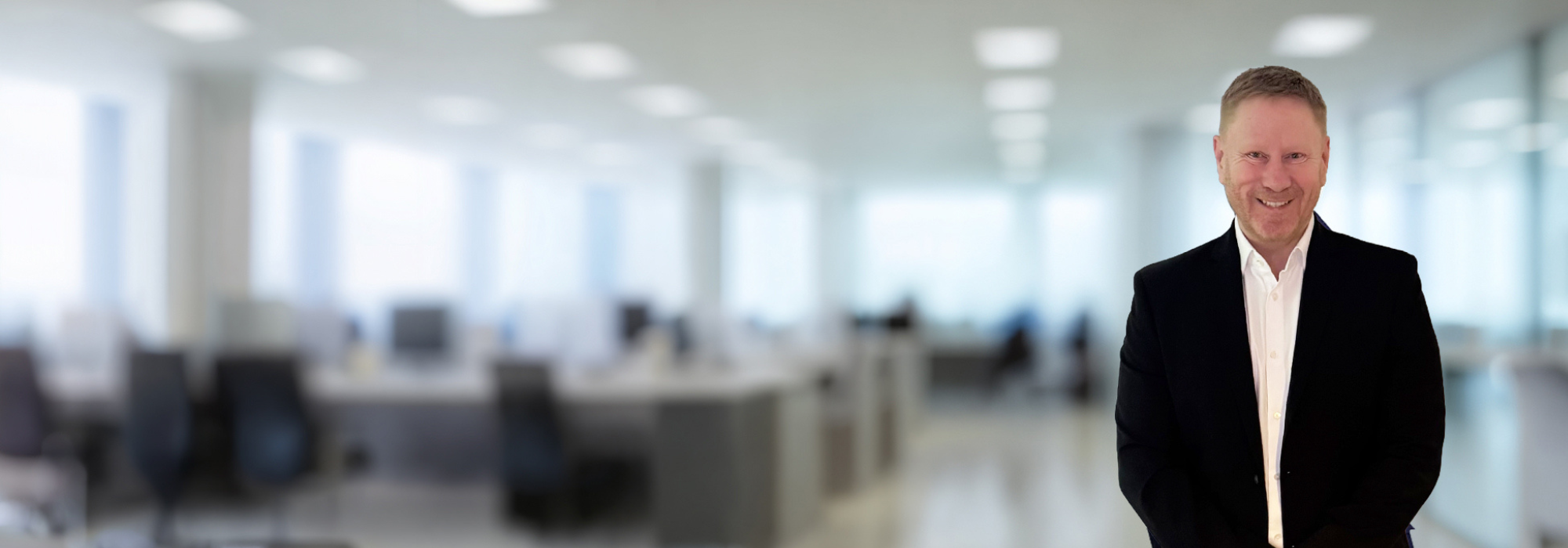 Smiling man with a black suit jacket and a white shirt. Office background behind with tables, chairs and monitors.