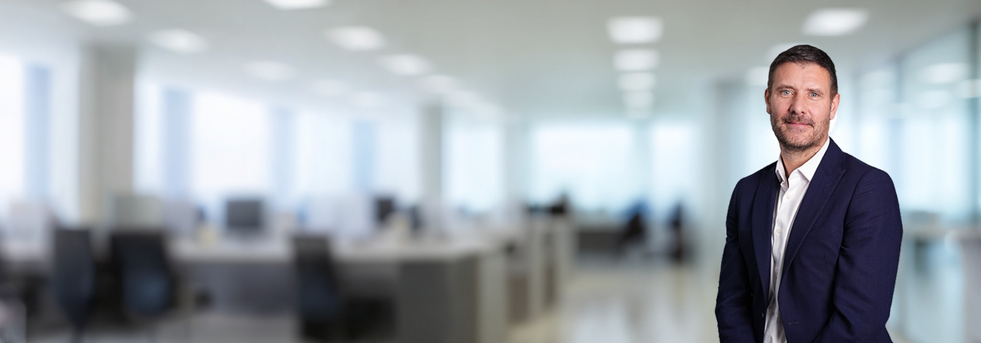 Smiling man with a blue blazer and a white shirt. Office desk and monitor behind. 