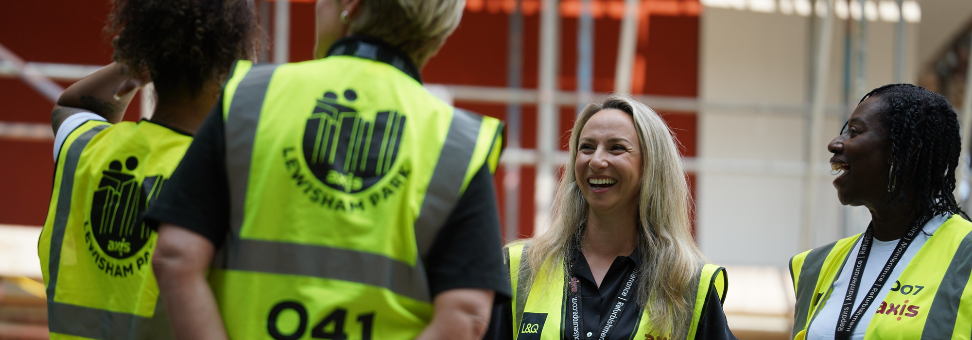 4 ladies: 2 from behind wearing hi-vis yellow jackets and 2 standing in front, smiling and laughing. Construction works behind. 