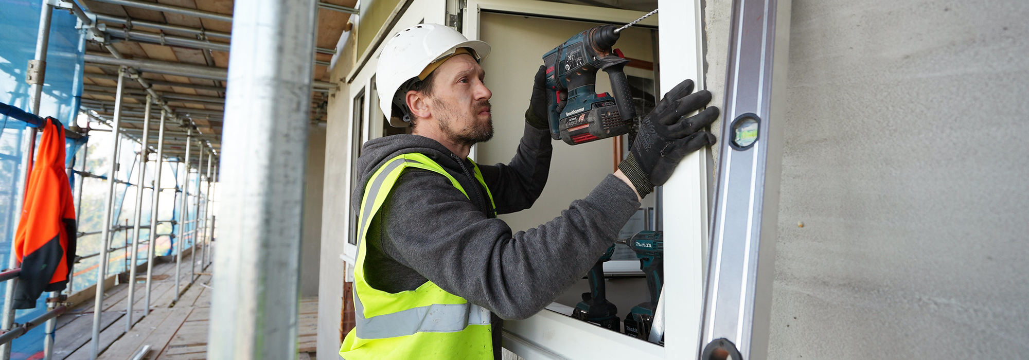 operative in high vis and hard hat using a drill to install window frame from exterior building scaffolding.