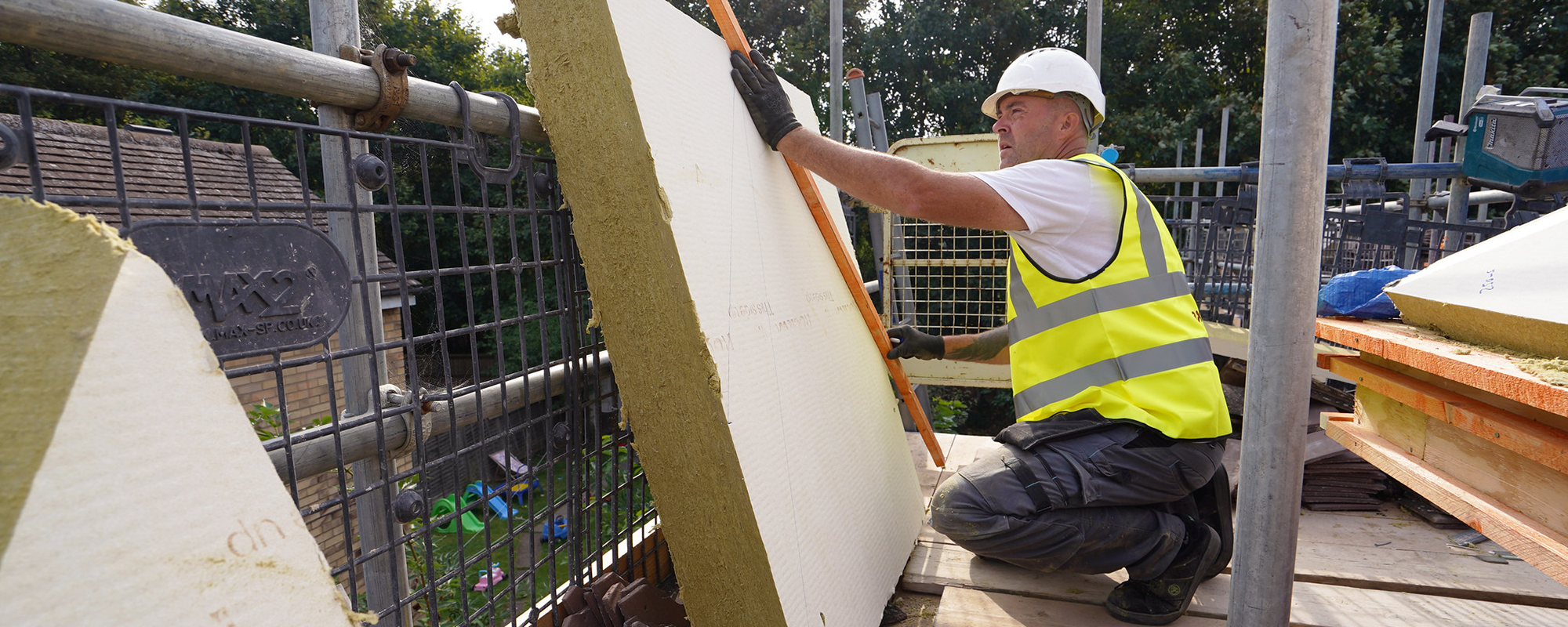 operative in high vis and a white hard hat measuring and cutting insulation on scaffolding