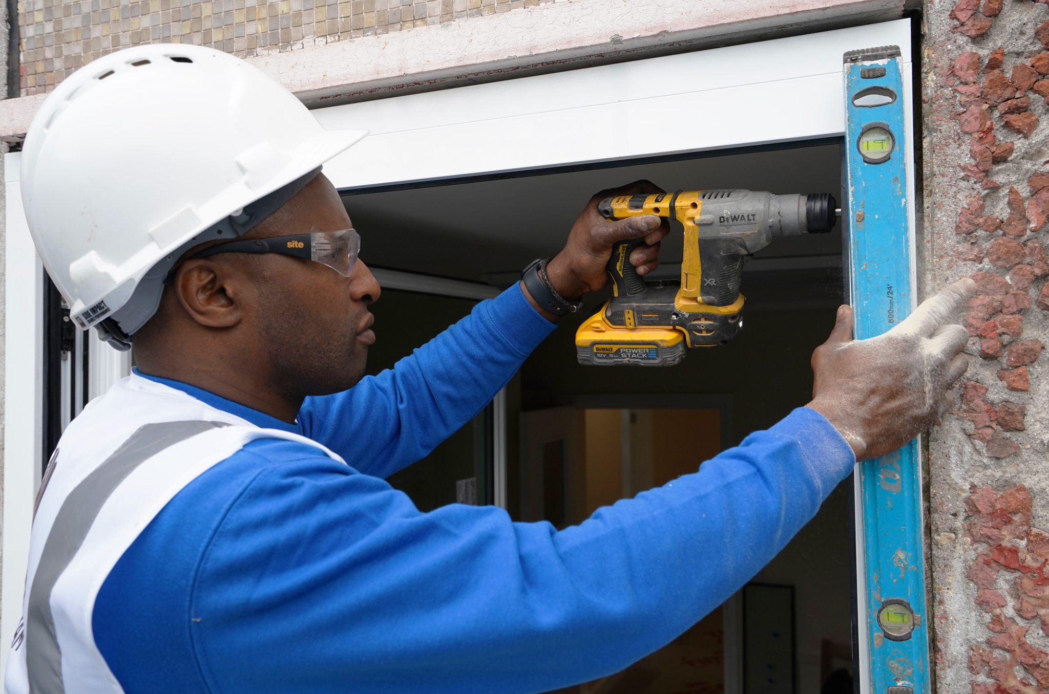 Man with white helmet, blue jumper and grey vest, holding a tool for the window frame