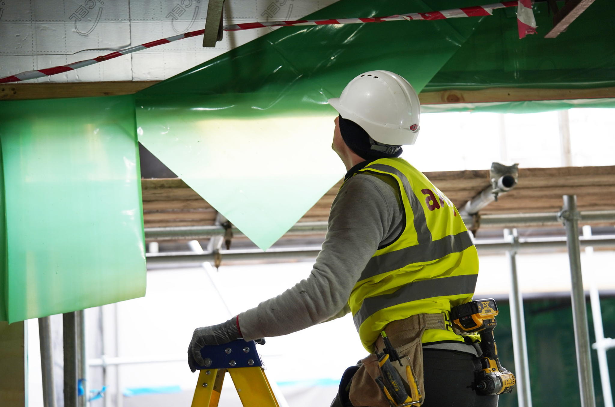 Man climbing up the top of a ladder, wearing a yellow vest, grey top, white helmet and gloves, looking at the ceiling