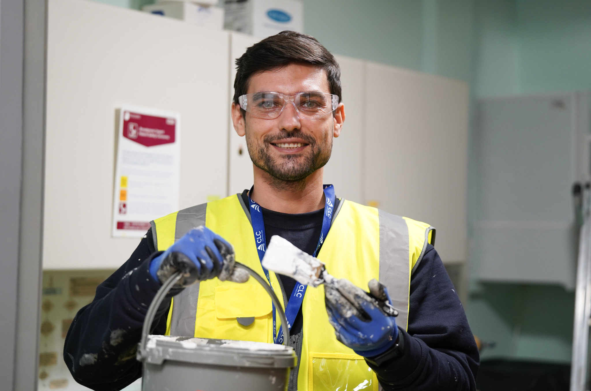 Man with glasses, paint brush and bucket of white paint. Wearing yellow vest, and blue top. Inside room with cupboards behind.
