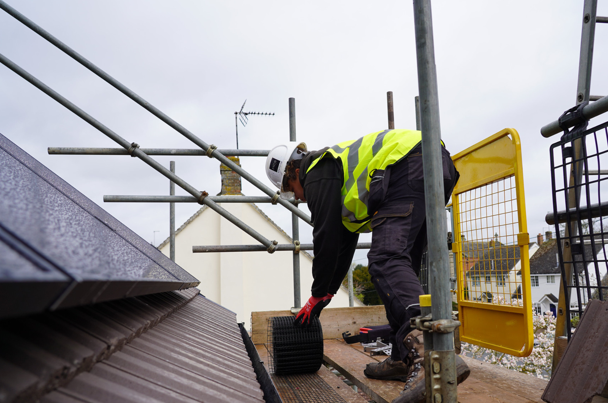 Woman wearing white helmet, yellow vest, black clothes and boot. On a roof with works.