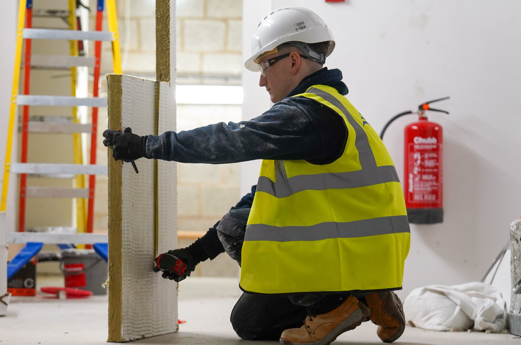Young boy with yellow vest, top and white helmet. Holding a wall with gloves. Ladder behind, in a white room