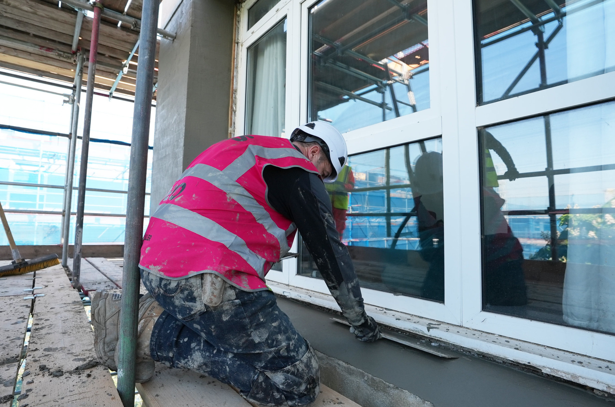 Man behind down fixing a window from the outside. Wearing purple vest, black clothes, white helmet and boots. Scaffolding all around.