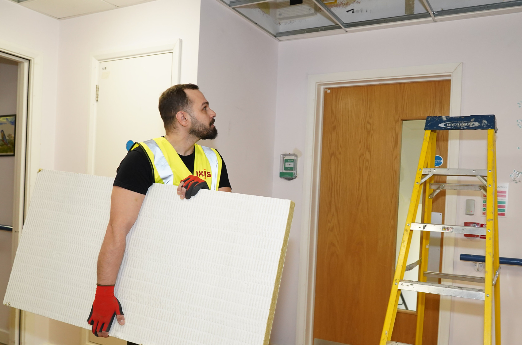 Man with a yellow vest, and black top, red gloves. Holding a white panel wall, looking up at the ceiling. Yellow ladder on the right. White walls, brown door.