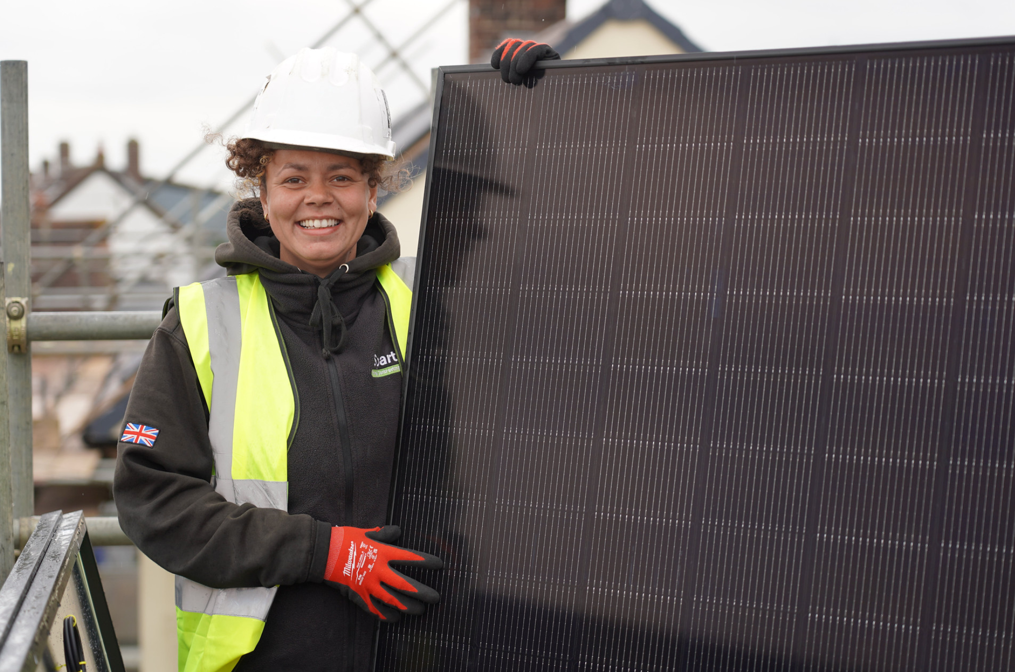 Woman holding a black solar panel. She's wearing a white helmet, black top, yellow vest with red gloves.