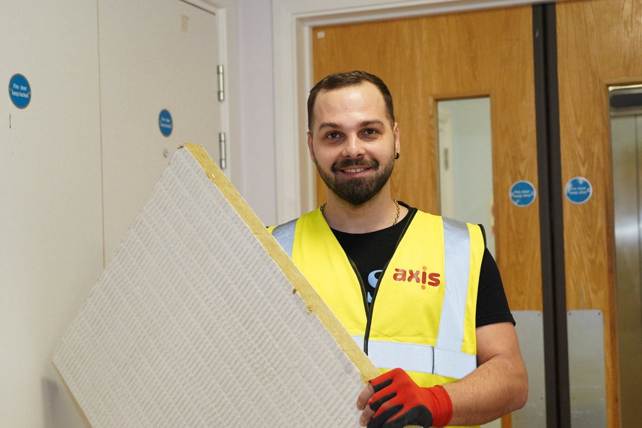 Man with yellow vest and axis logo holding a board. Fire doors behind and a white wall