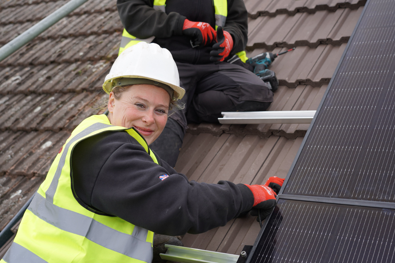 Woman with a yellow vest and a white helmet on a brown roof, smiling.
