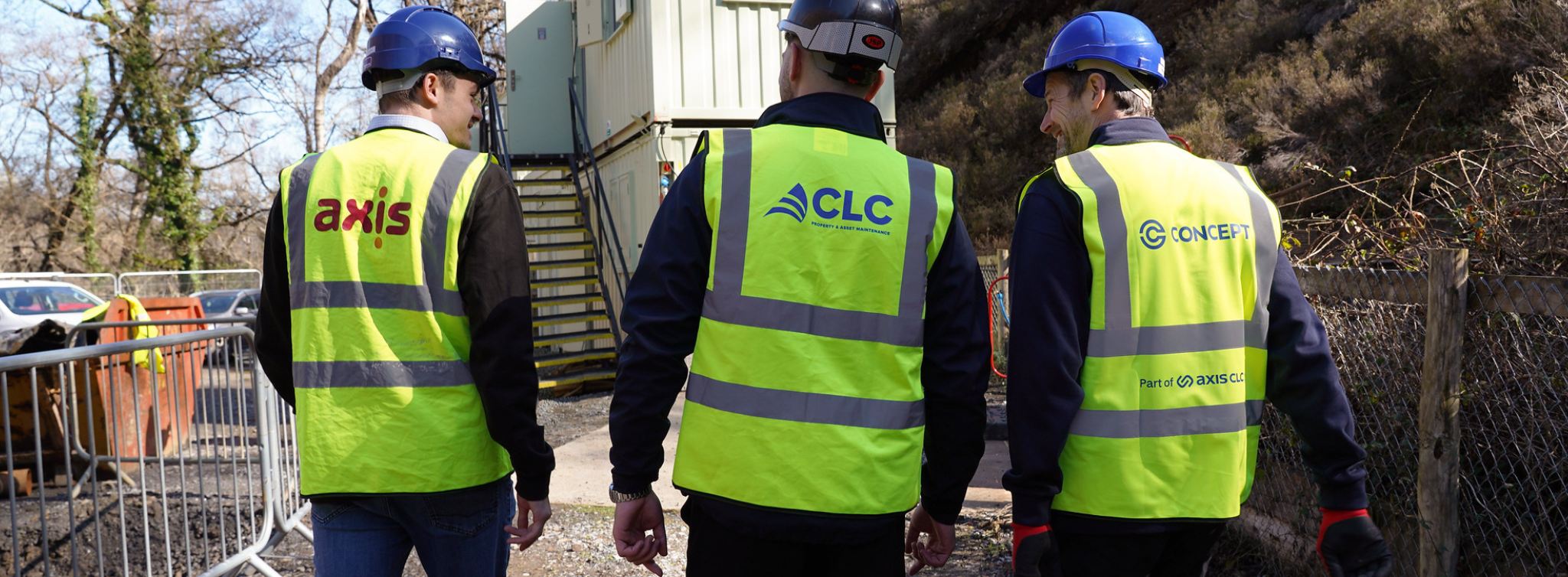 Three men wearing yellow vest and blue helmets. Logos on the back of the vest: Axis, CLC, Concept. Walking away outside