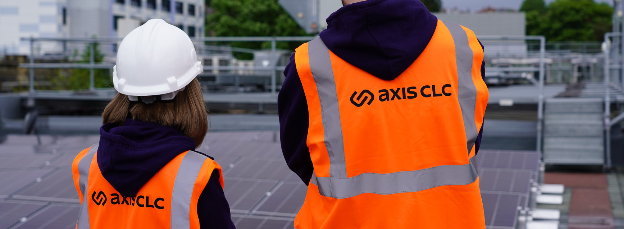 Two people with backs turned, white helmet, orange vets, logo with: axis clc on it. Outside.